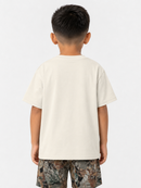 A child viewed from behind wearing a plain off-white short-sleeve t-shirt and camouflage shorts against a light gray studio background.