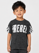 A young boy wearing a dark gray acid-wash t-shirt with a distressed white 'REBEL' graphic across the chest, photographed against a light gray studio background.