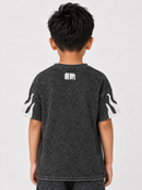 A child viewed from the back wearing a dark washed short-sleeve t-shirt with white graphic details on the sleeves and a white graphic print on the upper back, set against a plain light gray background.