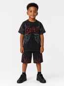A young boy modeling a black acid-wash graphic t-shirt and matching shorts set with red flame and gothic lettering details, paired with black sneakers, against a light gray studio background.