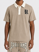 A man wearing a beige short-sleeve polo shirt with a cream collar and a black gothic letter 'R' on the chest, paired with matching beige shorts featuring a large 'SPARTANS' graphic.