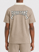 A person shown from the back wearing a beige short-sleeve polo shirt with a white collar and a large white 'SPARTANS' graphic across the upper back, paired with matching beige shorts against a light gray studio background.