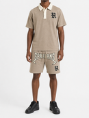 A model wearing a beige short-sleeve polo shirt and matching shorts set. The polo features a cream collar and a black embroidered letter 'R' on the chest.