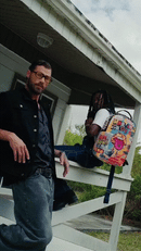 A colorful, highly detailed cartoon backpack with a blue strap is shown in a lifestyle setting, sitting on the porch of a white house next to two men.