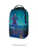 A sprayground backpack featuring a vibrant graphic print of the Joker standing on a car with graffiti-style text, set against a blue and green background. The backpack has a black top handle and zipper, and a Batman x Springboard collaboration logo is visible at the bottom.