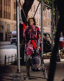 A model stands on a city sidewalk wearing a vibrant, multicolored patterned tunic top and matching zebra-print leggings, holding a black backpack with a red and white graphic design.