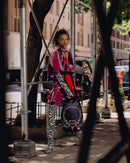 A model stands on a city sidewalk under scaffolding, wearing a vibrant magenta and purple patterned jacket, bold black-and-white zebra-print pants and arm warmers, and carrying a dark blue leopard-print shoulder bag.