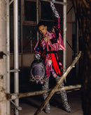 A model poses on industrial scaffolding wearing a vibrant pink, purple, and red patterned long-sleeve top, matching patterned thigh-high socks, and black-and-white zebra-print boots, while holding a dark purple animal-print backpack.