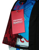 Close-up of a backpack with a bright blue and red graphic print, black straps, and a red Sprayground Monopoly hangtag attached.