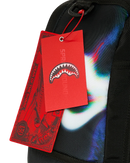 Close-up of a black backpack with a colorful abstract graphic print in blue, white, and green, featuring a red hangtag with a shark mouth logo and the brand name SPRAYGROUND.