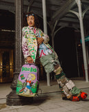 A person leans against a large column in an industrial covered walkway, wearing a heavily styled outfit with a colorful graffiti-print jacket, patchwork pants, and bright orange shoes, while holding a purple sprayground backpack featuring a cartoon character and the text 'Richie Rich'.