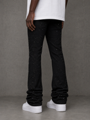 Black flare jeans with a subtle speckled texture, worn by a model with a white t-shirt and white platform sneakers against a gray concrete wall.