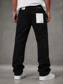 A person shown from the back wearing black jeans with white sneakers and a white t-shirt, standing against a gray concrete wall. The jeans have white paper labels attached to the back pockets.
