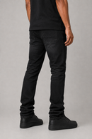 Rear three-quarter view of a person wearing slim-fit black jeans, a black t-shirt, and black sneakers against a neutral gray studio background.
