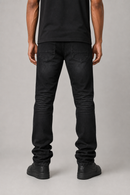 Rear view of a person wearing slim-fit black jeans, a black t-shirt, and black boots against a neutral gray studio background.