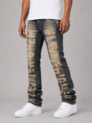 A person wearing slim-fit dark gray jeans with large distressed beige text running vertically down the front, paired with a white t-shirt and white sneakers, against a light gray studio background.